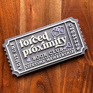 May include: A gray and white ticket-shaped item with the words "forced proximity" and "book club" printed on it. The ticket also includes the words "lifetime membership" and a serial number. The ticket has a key and pretzel design.