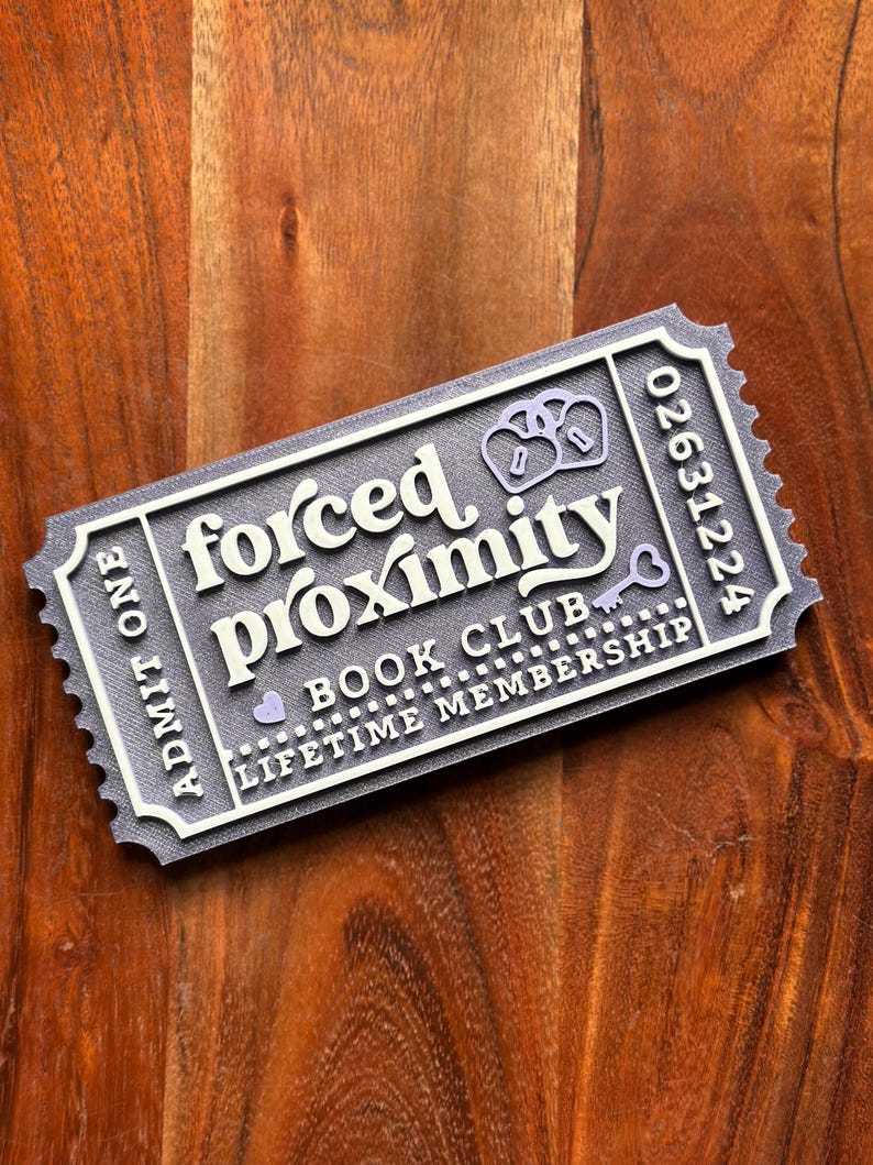 May include: A gray and white ticket-shaped item with the words "forced proximity" and "book club" printed on it. The ticket also includes the words "lifetime membership" and a serial number. The ticket has a key and pretzel design.
