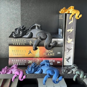 May include: A collection of dragon-shaped bookends and bookmarks in various colors, resting on and around a stack of books. The books have titles like "Rebecca Yarros" and "Iron Flame". The dragon figures are in black, gold, blue, purple, and green.