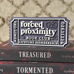 May include: A dark blue and white ticket-shaped sign that reads "forced proximity BOOK CLUB LIFETIME MEMBERSHIP" sits atop a stack of books. The books have red and white text that reads "TREASURED" and "TORMENTED".