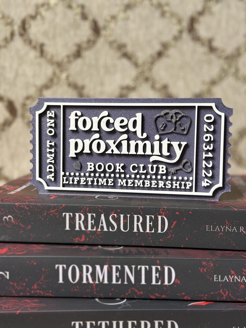 May include: A dark blue and white ticket-shaped sign that reads "forced proximity BOOK CLUB LIFETIME MEMBERSHIP" sits atop a stack of books. The books have red and white text that reads "TREASURED" and "TORMENTED".