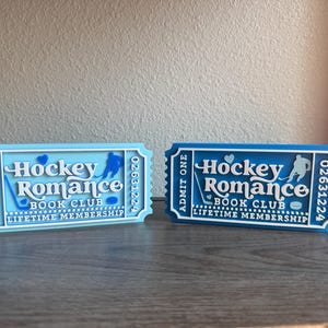 May include: Two decorative ticket-shaped signs. One is light blue, the other is dark blue. Both read "Hockey Romance Book Club Lifetime Membership." Each has a hockey player silhouette and the number 02631224.