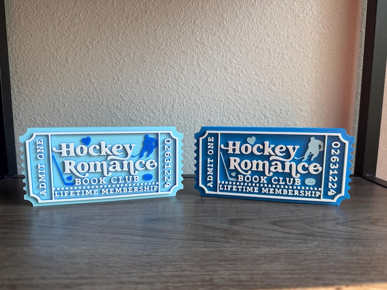 May include: Two decorative ticket-shaped signs. One is light blue, the other is dark blue. Both read "Hockey Romance Book Club Lifetime Membership." Each has a hockey player silhouette and the number 02631224.