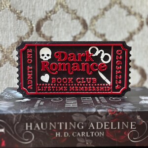 Puede incluir: Un objeto negro y rojo con forma de boleto con las palabras "Dark Romance Book Club Lifetime Membership" y una calavera, esposas y un cuchillo. El boleto est&aacute; colocado encima de un libro titulado "Haunting Adeline" de H.D. Carlton.