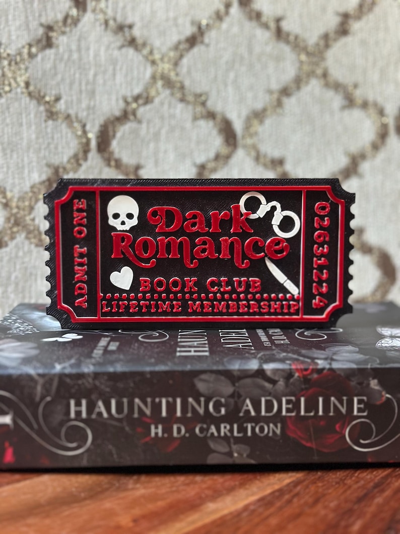 Puede incluir: Un objeto negro y rojo con forma de boleto con las palabras "Dark Romance Book Club Lifetime Membership" y una calavera, esposas y un cuchillo. El boleto est&aacute; colocado encima de un libro titulado "Haunting Adeline" de H.D. Carlton.