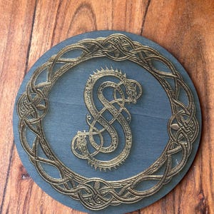 May include: A round wooden coaster with a dark blue background and a gold-colored Celtic knot design. The center of the coaster features a gold-colored dragon design.