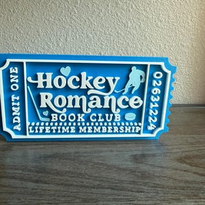 May include: A blue and white 3D-printed ticket with the words "Hockey Romance Book Club" and a hockey player silhouette. The ticket reads "Admit One" and "Lifetime Membership". A heart and hockey stick are also featured.
