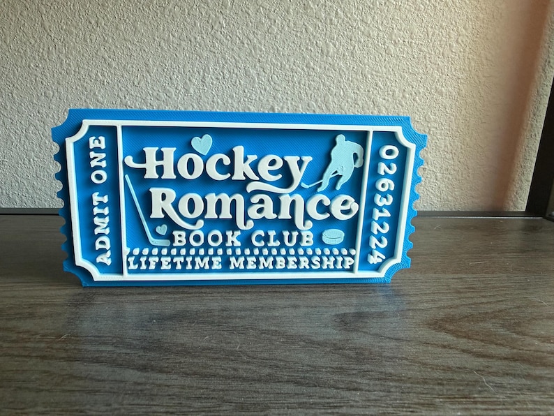 May include: A blue and white 3D-printed ticket with the words "Hockey Romance Book Club" and a hockey player silhouette. The ticket reads "Admit One" and "Lifetime Membership". A heart and hockey stick are also featured.