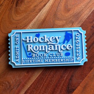 May include: A light blue and white layered ticket-shaped sign with the words "Hockey Romance Book Club Lifetime Membership." The sign features a hockey player silhouette, a hockey stick, and a puck.