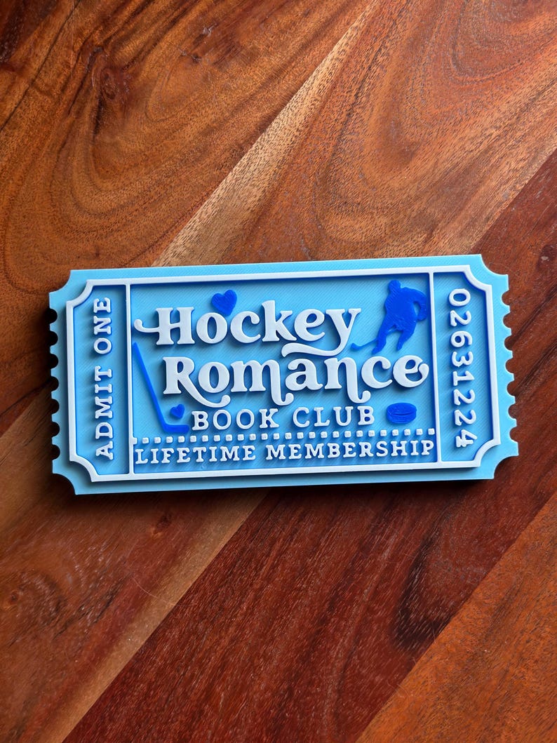 May include: A light blue and white layered ticket-shaped sign with the words "Hockey Romance Book Club Lifetime Membership." The sign features a hockey player silhouette, a hockey stick, and a puck.