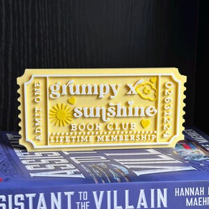 Puede incluir: Un boleto amarillo y blanco impreso en 3D con el texto "grumpy x sunshine BOOK CLUB LIFETIME MEMBERSHIP". El boleto tiene un dise&ntilde;o de sol y coraz&oacute;n. El boleto est&aacute; colocado encima de un libro.