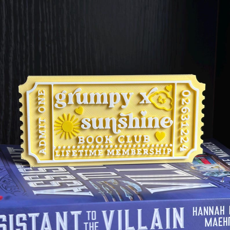 Puede incluir: Un boleto amarillo y blanco impreso en 3D con el texto "grumpy x sunshine BOOK CLUB LIFETIME MEMBERSHIP". El boleto tiene un dise&ntilde;o de sol y coraz&oacute;n. El boleto est&aacute; colocado encima de un libro.