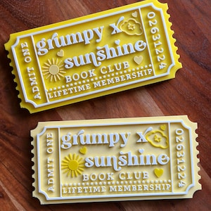 Puede incluir: Dos boletos amarillos y blancos con el texto "grumpy x sunshine BOOK CLUB LIFETIME MEMBERSHIP". Los boletos tienen un dise&ntilde;o de sol, nube y coraz&oacute;n. Los boletos son rectangulares con un borde festoneado y las palabras "ADMIT ONE" a la izquierda.