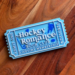 May include: A light blue and white ticket-shaped item with the words "Hockey Romance Book Club Lifetime Membership". The ticket has a hockey player silhouette, a hockey stick, and a puck. The ticket also has the number "02631224".