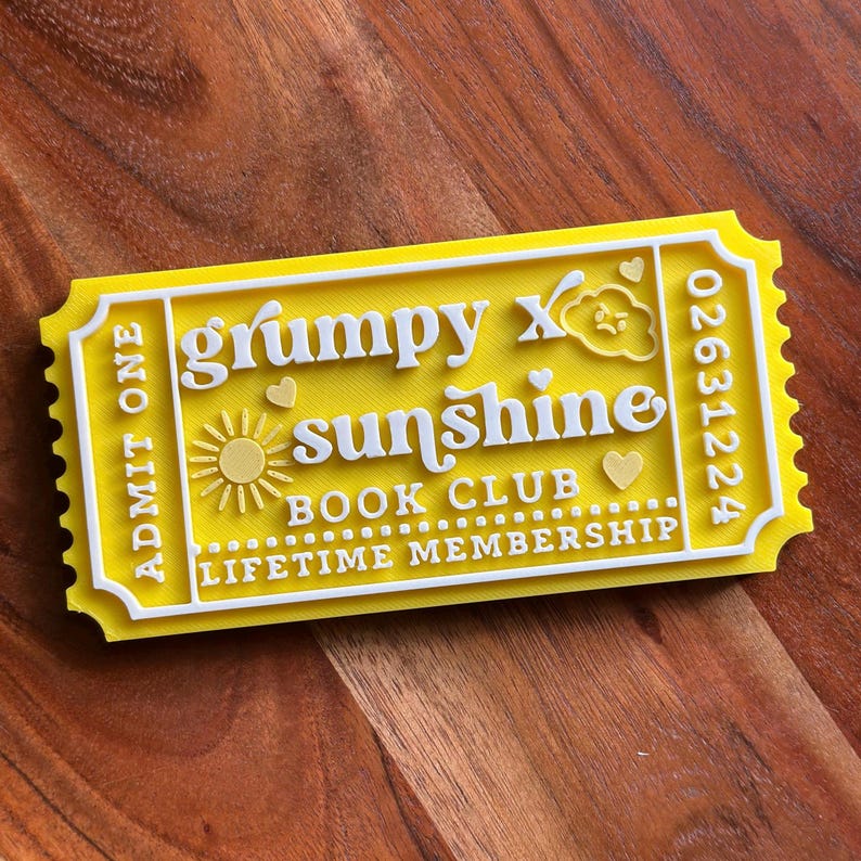 Puede incluir: Un ticket amarillo con letras blancas, con las palabras "grumpy x sunshine book club lifetime membership". El dise&ntilde;o incluye un sol y corazones. El ticket tambi&eacute;n tiene las palabras "Admit One" y un n&uacute;mero.