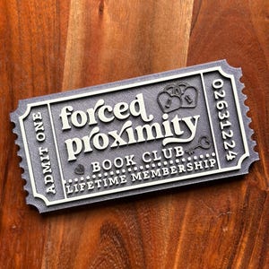 May include: A gray ticket-shaped sign with the words "forced proximity" in white, along with "book club" and "lifetime membership." The sign has a decorative border and the number "02631224." The background is a wooden surface.