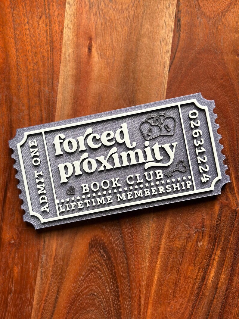 May include: A gray ticket-shaped sign with the words "forced proximity" in white, along with "book club" and "lifetime membership." The sign has a decorative border and the number "02631224." The background is a wooden surface.
