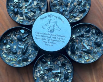 May include: Five black candles with skull accents and a variety of stones and herbs. The candles are in black tins and are arranged on a wooden surface. The center candle has a label that reads "Hades Offering Candle."