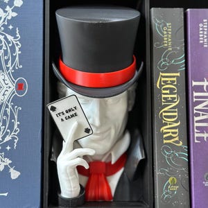 May include: A magician bust with a black top hat, red band, and red bow tie, holding a playing card that reads "IT'S ONLY A GAME." The bust is displayed in a black shelf with playing cards in front and books on the right.