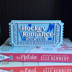May include: A light blue ticket-shaped bookmark with the words "Hockey Romance Book Club Lifetime Membership" and a hockey player silhouette. The bookmark sits atop two books by Elle Kennedy, titled "The Mistake" and "The Deal".