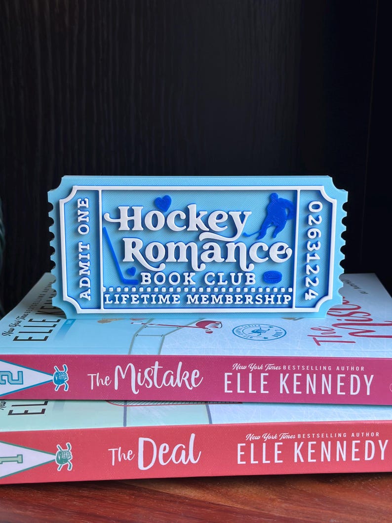 May include: A light blue ticket-shaped bookmark with the words "Hockey Romance Book Club Lifetime Membership" and a hockey player silhouette. The bookmark sits atop two books by Elle Kennedy, titled "The Mistake" and "The Deal".