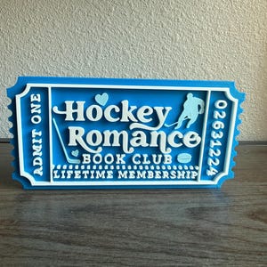 May include: A blue and white ticket-shaped sign that reads "Hockey Romance Book Club Lifetime Membership." The sign features a hockey player silhouette, a hockey stick, a puck, and two hearts. The text "Admit One" is on the left side.
