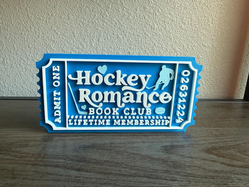 May include: A blue and white ticket-shaped sign that reads "Hockey Romance Book Club Lifetime Membership." The sign features a hockey player silhouette, a hockey stick, a puck, and two hearts. The text "Admit One" is on the left side.