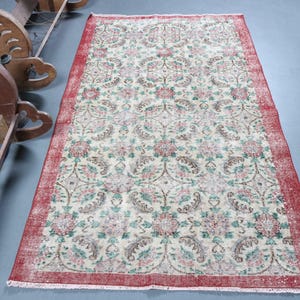 May include: A rectangular, vintage-style rug with a cream-colored field and a red border. The rug features a floral pattern in shades of red, green, and brown. The rug is placed on a gray surface, and a wooden rack is visible on the left.