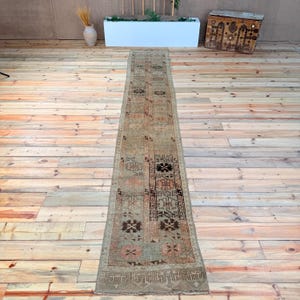 Op de afbeelding: Een lang, smal loperkleed met een vervaagd, gedempt kleurenpalet van beige, zachtroze en donkerbruin. Het kleed toont een reeks herhalende geometrische en florale motieven gerangschikt in verticale panelen, afgewerkt met een decoratieve rand.