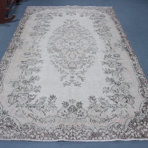 May include: A rectangular area rug featuring a cream-colored field with a central dark brown floral medallion. The border displays a dense, intricate pattern of dark brown botanical motifs and scrolling vines against a lighter background.