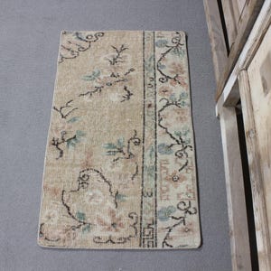 Peut inclure: Tapis rectangulaire beige avec des motifs floraux verts, roses et noirs. Le tapis a un aspect vieilli et vintage, avec un motif de bordure sur un côté. Le tapis est posé sur une surface grise.