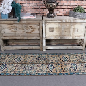 May include: A vintage style runner rug with a cream background and a blue and brown floral pattern. The rug is placed in front of two wooden cabinets with a decorative metal teapot on top of one of the cabinets.