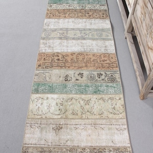 Pode incluir: Um tapete longo e retangular com um design de patchwork. O tapete é feito de tiras de tecido em vários tons de bege, marrom e verde. O tecido tem um visual vintage com cores desbotadas e padrões intrincados.