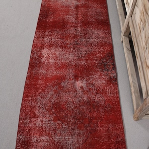 Könnte beinhalten: Ein langer, rechteckiger roter Teppich mit einem Used-Look im Vintage-Stil. Der Teppich hat ein verblasstes, gesprenkeltes Muster mit helleren Bereichen und dunkleren Akzenten, wodurch ein strukturierter Look entsteht. Die Kanten des Teppichs sind leicht ausgefranst.