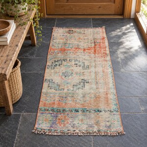 May include: A rectangular runner rug featuring a distressed pattern in shades of orange, teal, and cream. The rug has a woven texture with a repeating geometric motif and short fringe detailing along the edges, laid flat on a dark stone floor.