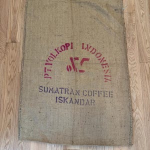 Könnte beinhalten: Ein Kaffeesack aus Jute mit rotem Text, der "P.T. YOLKOPI INDONESIA" in einem Bogen und "SUMATRAN COFFEE ISKANDAR" darunter zeigt. Der Sack ist rechteckig und hat ein raues, strukturiertes Aussehen.