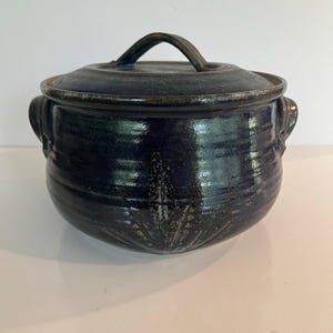 May include: A round, dark blue ceramic pot with a matching lid and two side handles. The exterior features a glossy, mottled glaze with a subtle, etched botanical pattern near the base. The pot has a wide, rounded body and a thick, sturdy rim.
