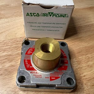 Peut inclure: Un pressostat ASCO Tri Point avec un raccord en laiton et un corps argenté. La boîte au-dessus de l'interrupteur indique "Pressure and Temperature Switches" en anglais, allemand et espagnol. Mesures en PSI et BAR.