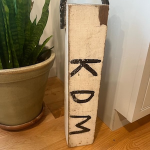 Puede incluir: Un letrero de madera hecho a mano con la palabra "Welcome" en un estilo manuscrito. El letrero pintado de blanco tiene un acabado envejecido y una pequeña hoja verde en la parte superior. Para uso interior o exterior.