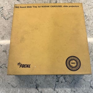 Puede incluir: Una bandeja de diapositivas Kodak Carousel vintage, modelo 140, en una caja cuadrada de cartón marrón. La caja tiene el texto "140 Round Slide Tray for KODAK CAROUSEL slide projectors" y el logotipo "FOCAL". Un gráfico circular con "140" está en la caja.