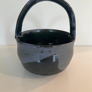 Puede incluir: Una cesta de cerámica hecha a mano con asa. La cesta es una mezcla de azul oscuro, gris y negro. El asa es azul oscuro y el interior es negro. El exterior tiene un aspecto moteado y texturizado.