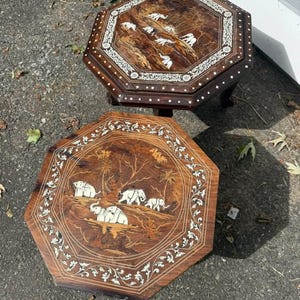 May include: Two octagonal wooden tables featuring intricate inlaid designs. The surfaces display detailed scenes of elephants in a natural landscape, surrounded by ornate floral borders. The wood grain is visible, and the inlay work is light-colored.
