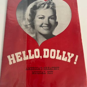 Può includere: Una copertina di album rossa con una foto in bianco e nero del volto di una donna all'interno di un cuore. Le parole "HELLO, DOLLY!" sono in bianco, con "AMERICA'S GREATEST MUSICAL HIT" sotto.