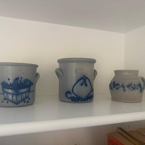 Puede incluir: Tres vasijas de cerámica gris con diseños pintados en azul. La vasija de la izquierda muestra una cesta de frutas, la central presenta un motivo en forma de corazón y la de la derecha tiene un patrón de enredadera. Tienen formas cilíndricas.