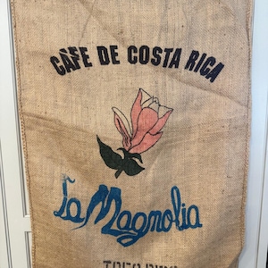 May include: A burlap coffee sack with the text "CAFE DE COSTA RICA" in black, a pink and green floral design, and the words "La Magnolia" and "TRES RIOS" in blue. The sack is hanging on a white surface.