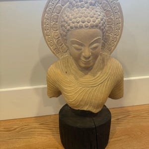 Può includere: Busto di Buddha beige con un'aureola dettagliata. La scultura è montata su una base di legno scuro e cilindrica. Il volto del Buddha è sereno e il busto è scolpito con dettagli intricati. Lo sfondo è una parete neutra e un pavimento in legno.