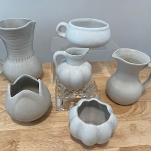May include: A collection of six off-white ceramic vessels. The assortment includes pitchers, a small cup, and decorative bowls. The pieces vary in shape and size, with some featuring textured surfaces and handles. The items are arranged on a wooden surface.