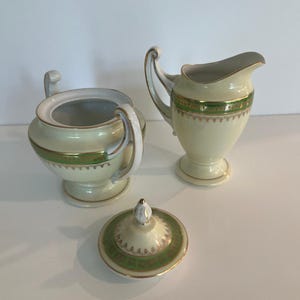 Puede incluir: Juego de azucarero y jarra de porcelana color crema con ribetes verdes y dorados. El azucarero tiene tapa y dos asas. La jarra tiene asa y pico. El juego está sobre una superficie blanca.