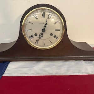 May include: A dark brown wooden mantel clock with a circular face, a gold-toned bezel, and black Roman numerals. The clock features black ornate hands and the Howard Miller brand name printed on the off-white dial, with two winding holes present.