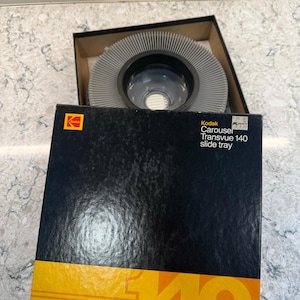 Puede incluir: Una bandeja de diapositivas Kodak Carousel Transvue 140 en su caja original. La caja es negra con una franja amarilla y el número 140. La bandeja es gris y circular, con un centro de plástico transparente.